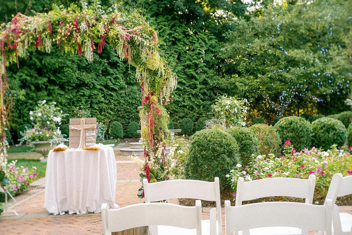 Ceremonia de boda en jard&iacute;n con arco floral decorado, sillas blancas y set de bienvenida